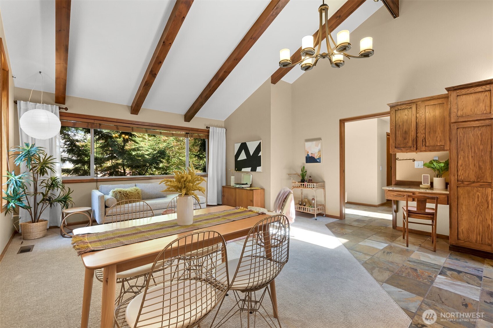 Dining and living area with exposed wood beams, a long table, and a large window overlooking trees.