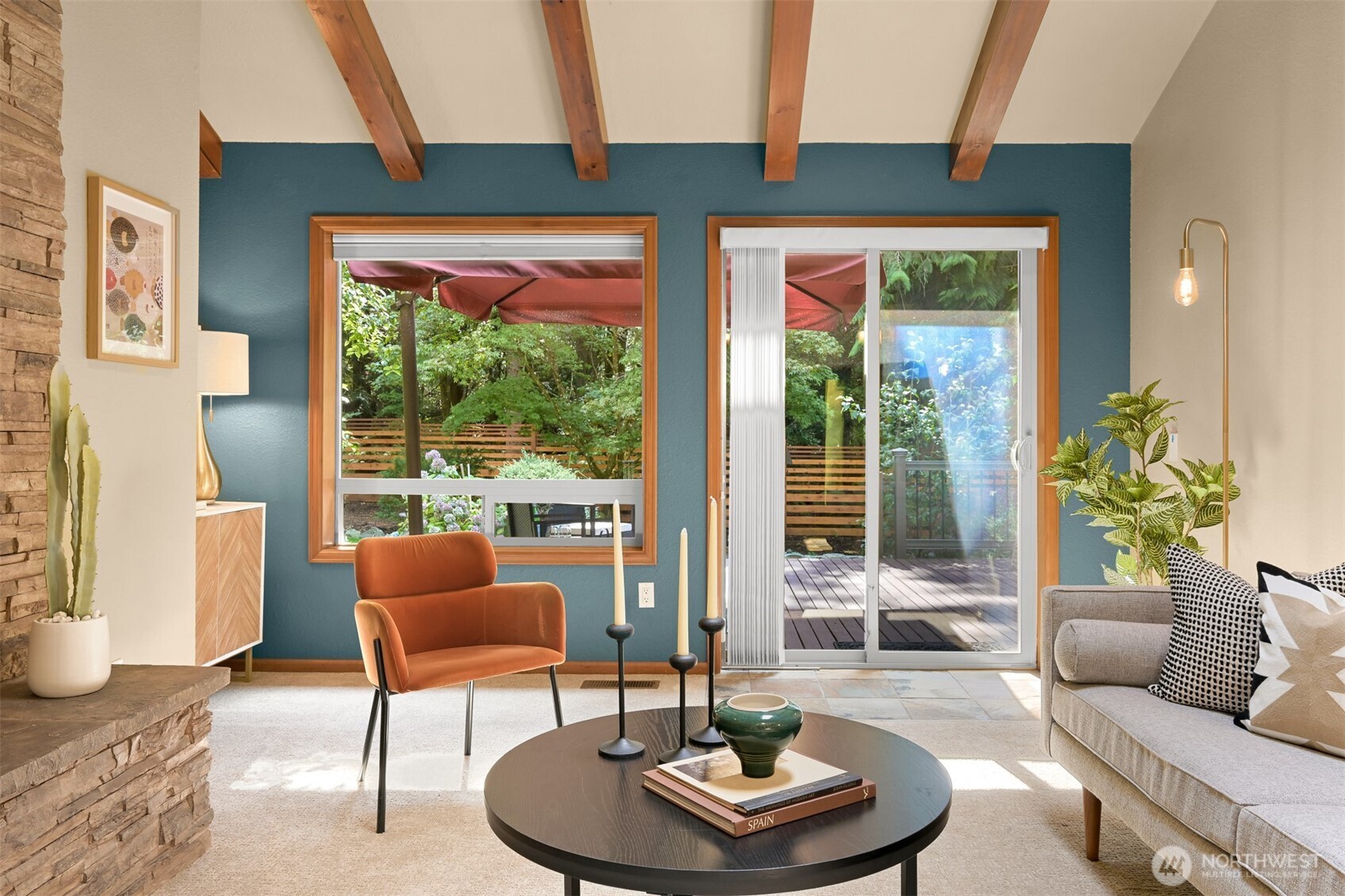 Living room with exposed beams, teal wall, stone fireplace, and sliding doors to a deck and leafy yard.