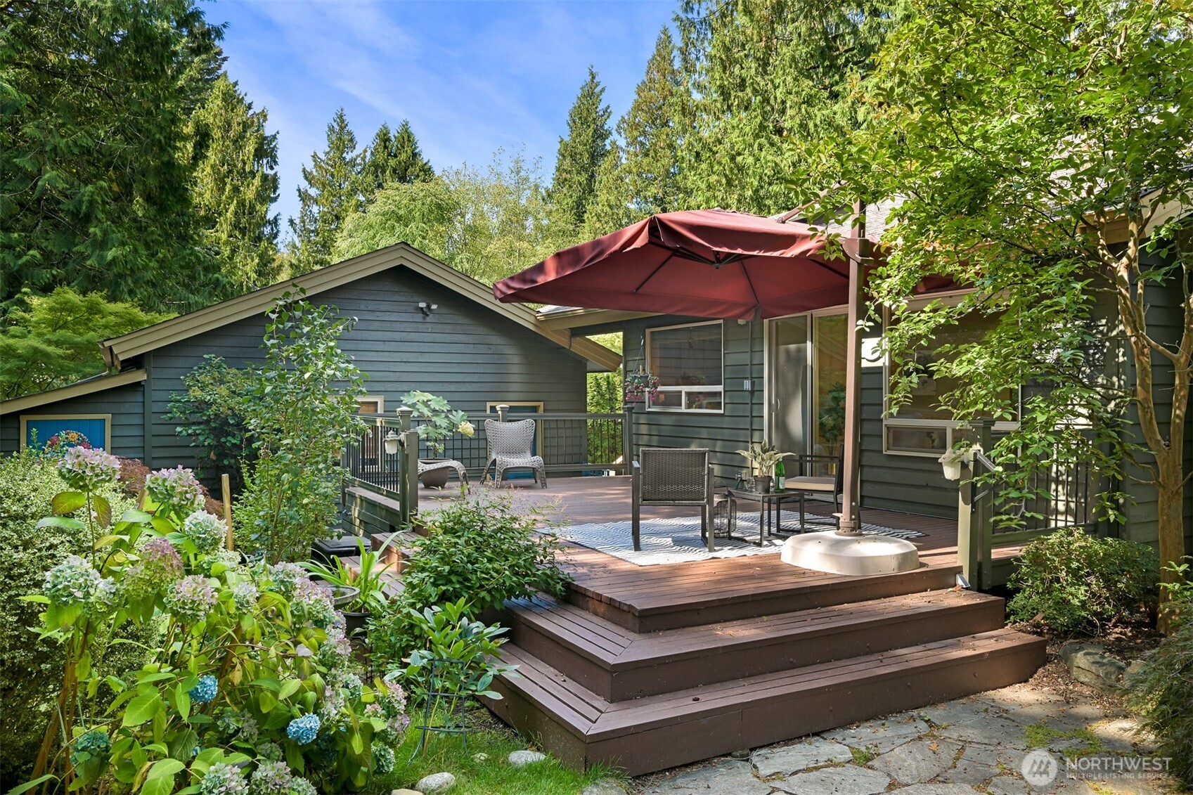 Raised deck with outdoor seating and a burgundy umbrella, surrounded by flowering hydrangeas and evergreens.