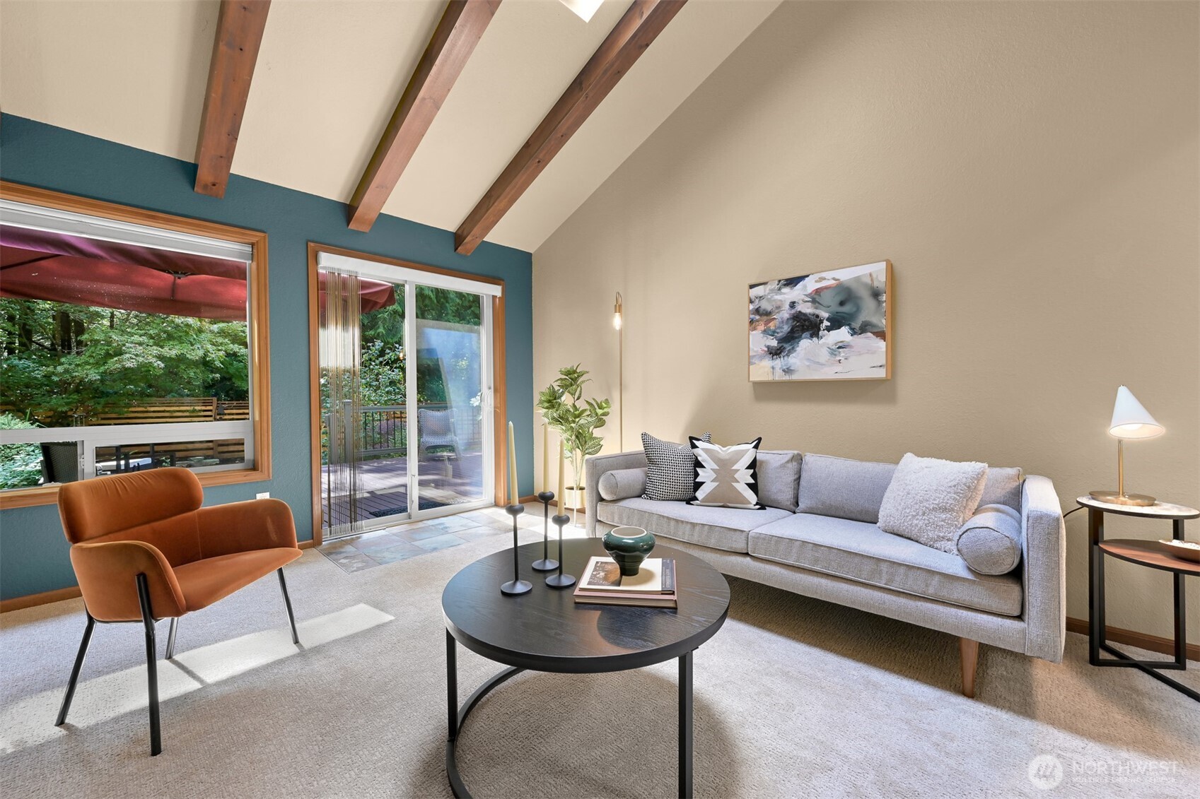 Staged living room with exposed wood beams, a teal accent wall, and windows overlooking a wooded deck.