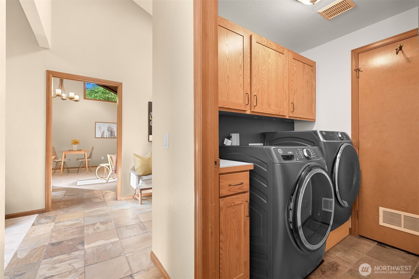 Laundry room with front-loading washer and dryer beneath wood cabinets, beside a tiled hallway.