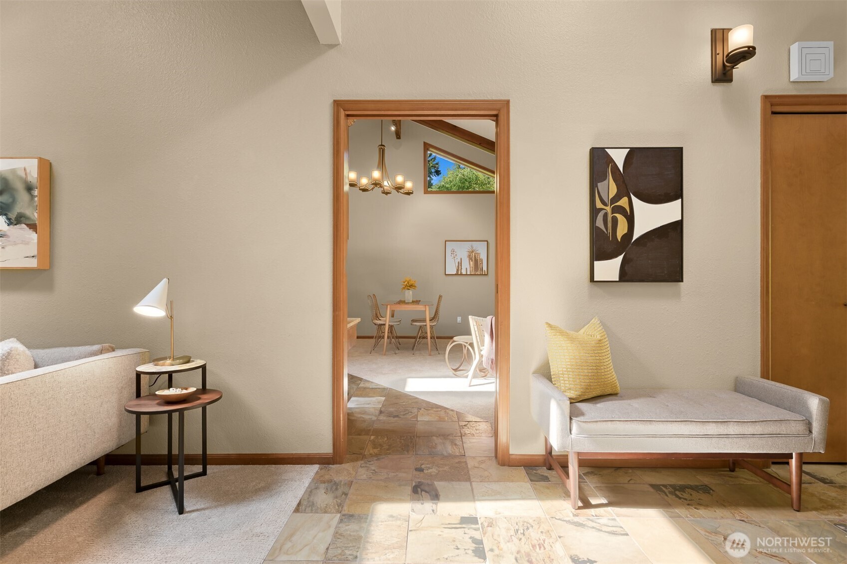 Staged living room with cream seating and stone tile, opening through a wood-trimmed doorway to a dining area.