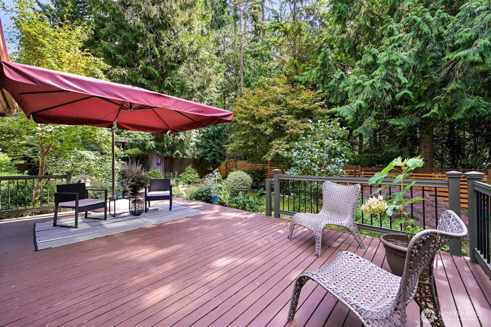 Wooden deck with wicker seating and a burgundy umbrella overlooking a lush garden.