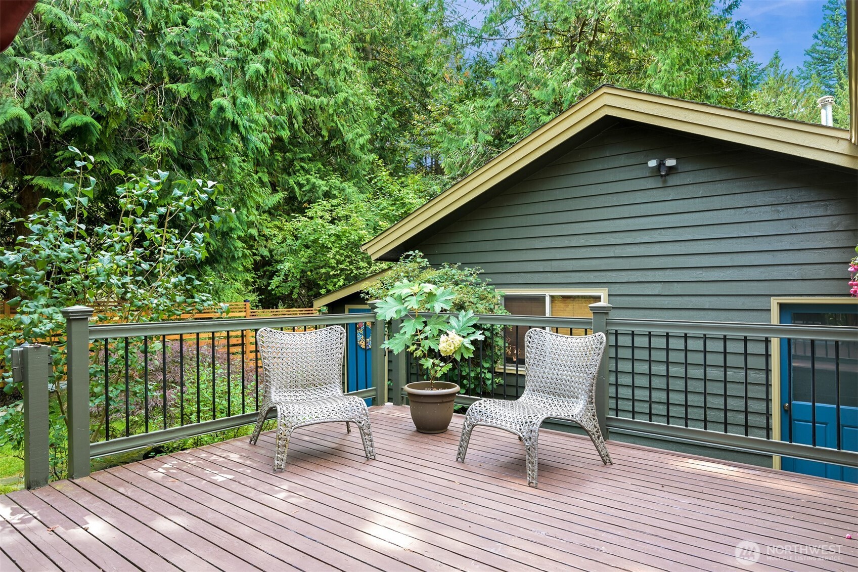 Wooden deck with two woven chairs and a potted plant beside a dark green house, surrounded by trees.