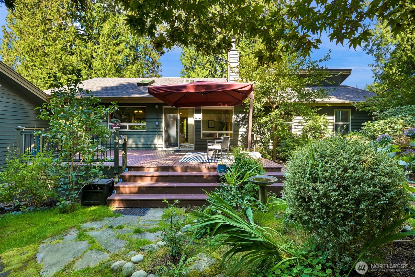 Home’s rear deck with a red umbrella, outdoor seating, and lush garden landscaping.