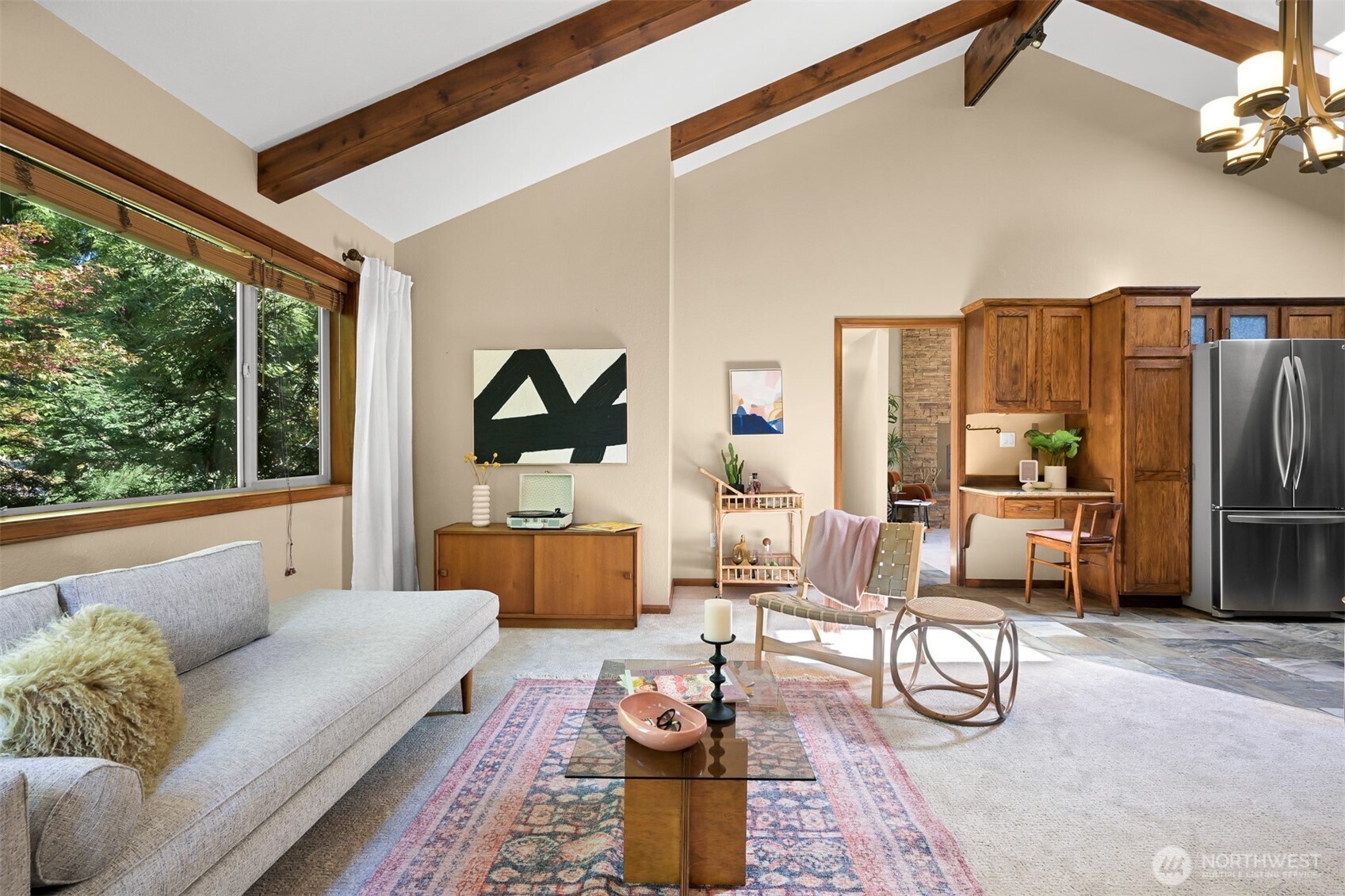 Living room with vaulted wood-beam ceiling, large tree-facing window, and open kitchen.