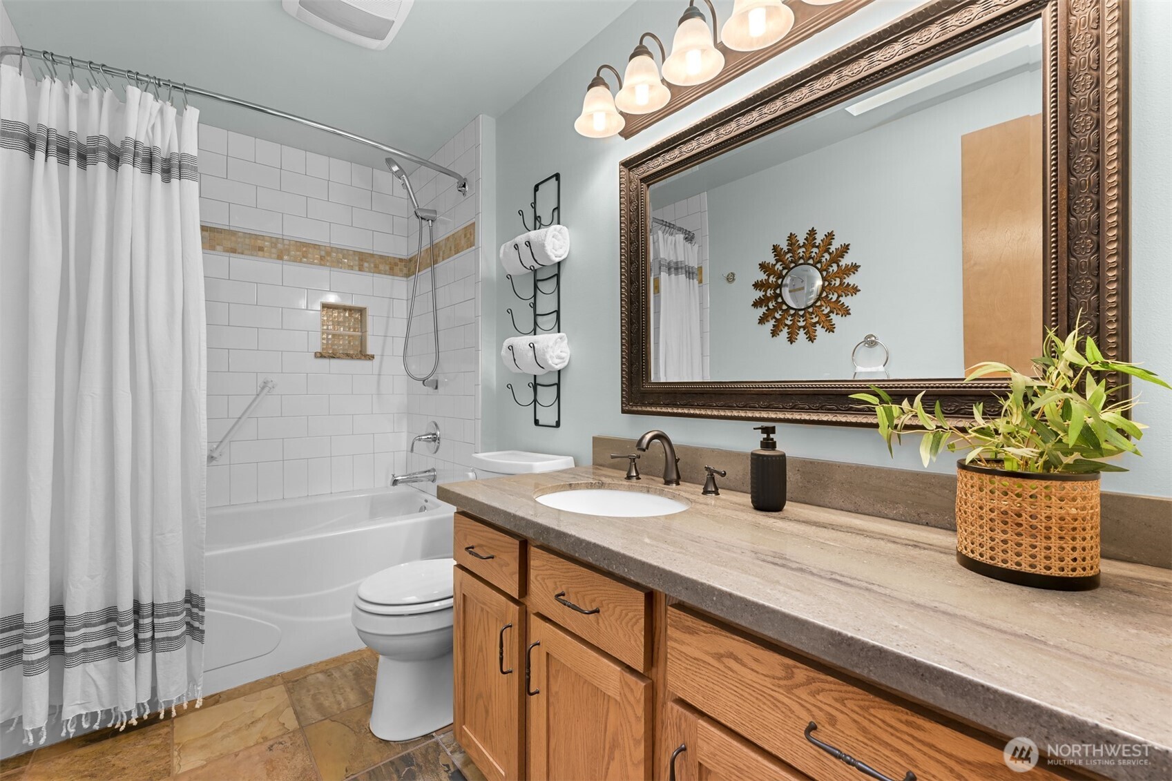 Bathroom with tiled tub and shower, long stone vanity, oak cabinets, and an ornate framed mirror.