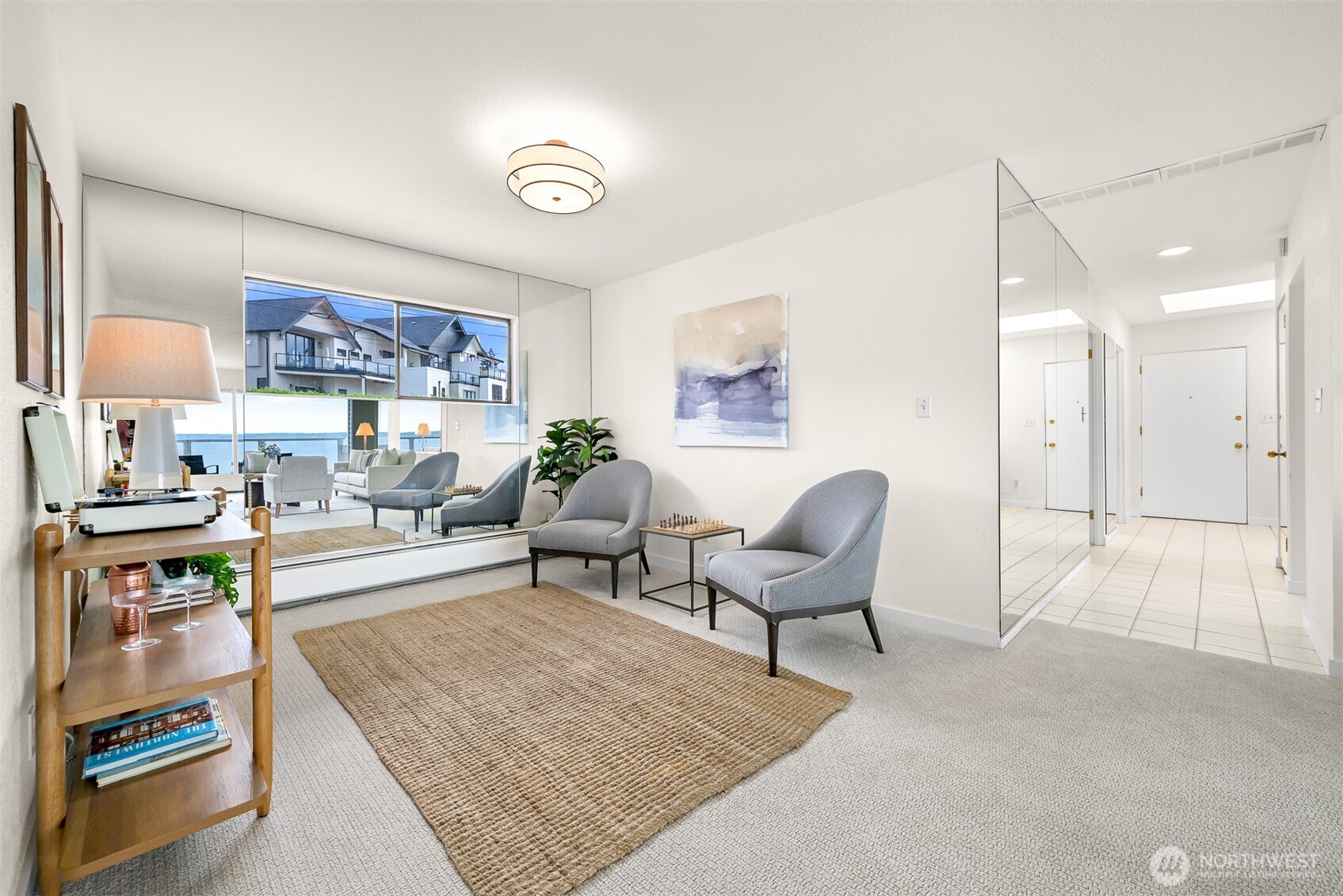 Living room with gray chairs, a woven rug, mirrored wall, and water visible through large windows.