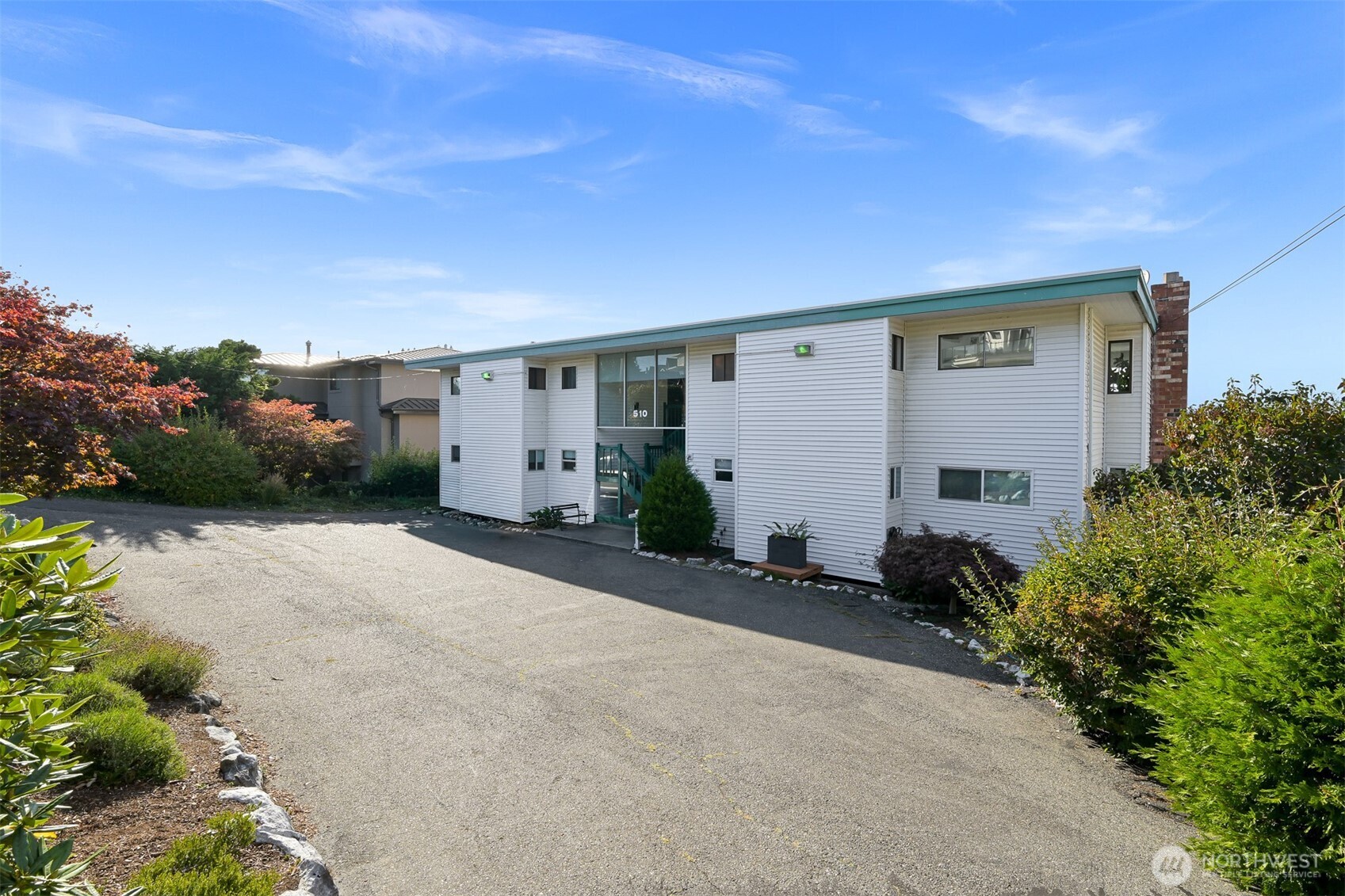 White multi-unit building with green roof trim, a paved parking area, and landscaped shrubs.
