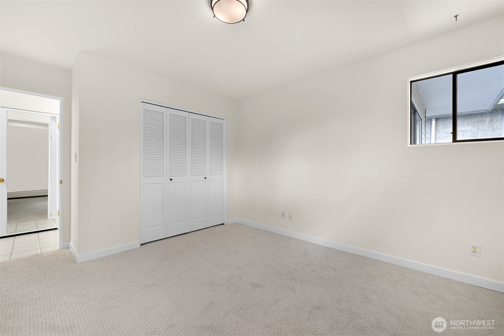 Empty bedroom with light carpet, louvered closet doors, and a window facing a nearby building.