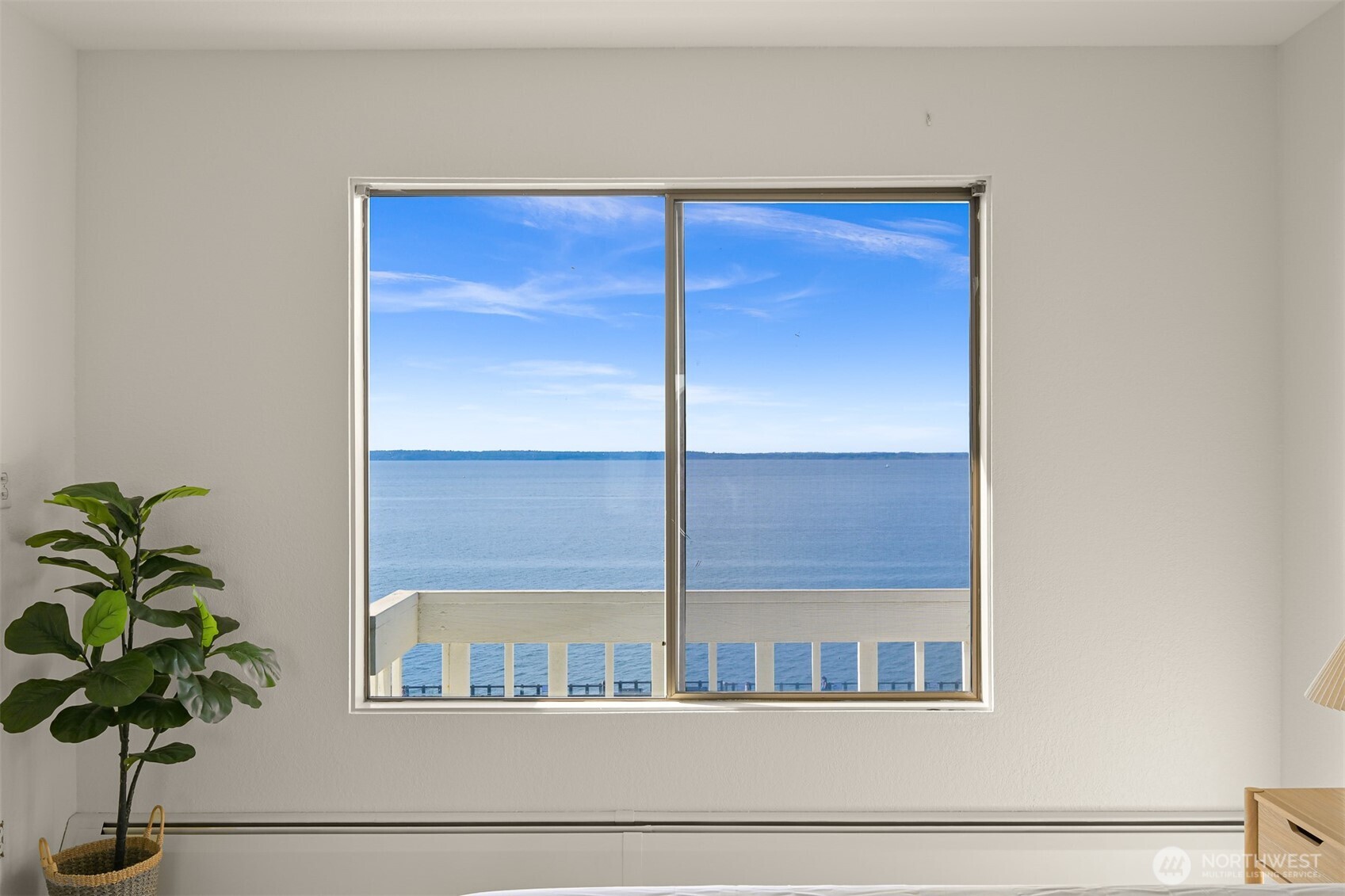 Room window overlooking calm blue water, with a white balcony railing and potted plant.