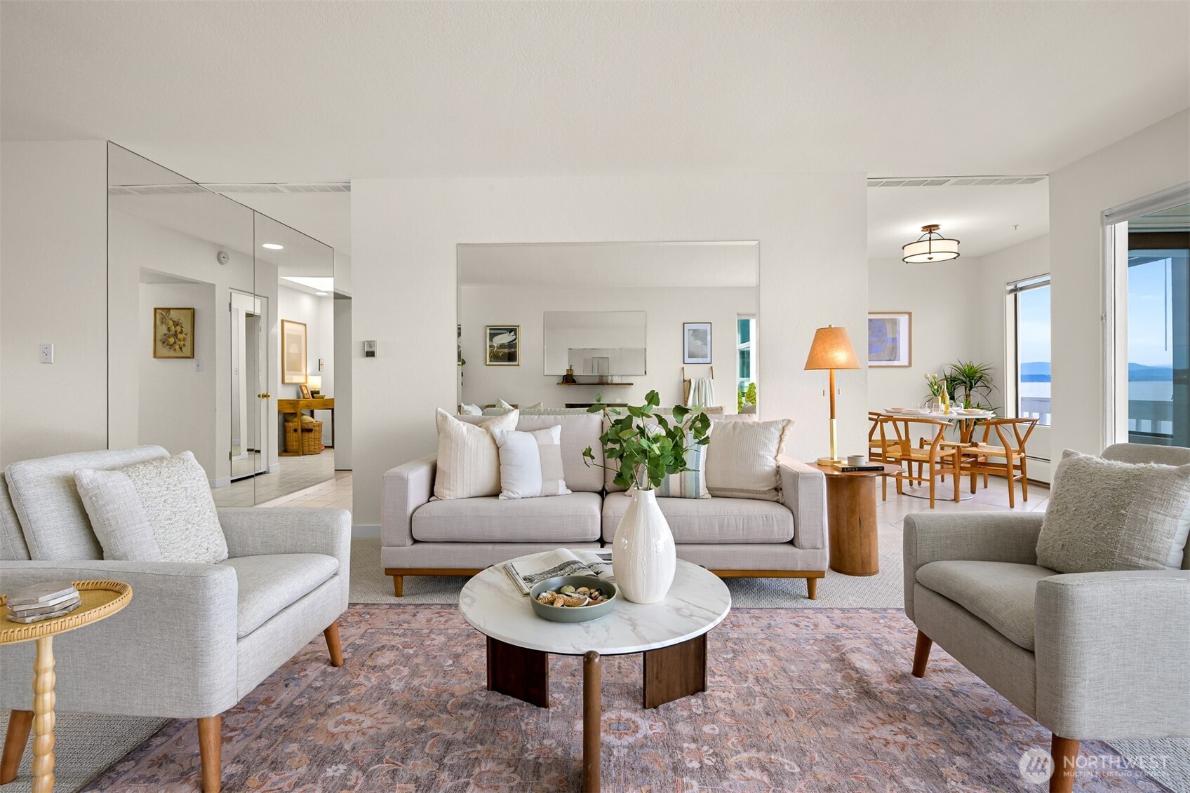 Furnished living room with pale sofas and a dining area overlooking water through large windows.