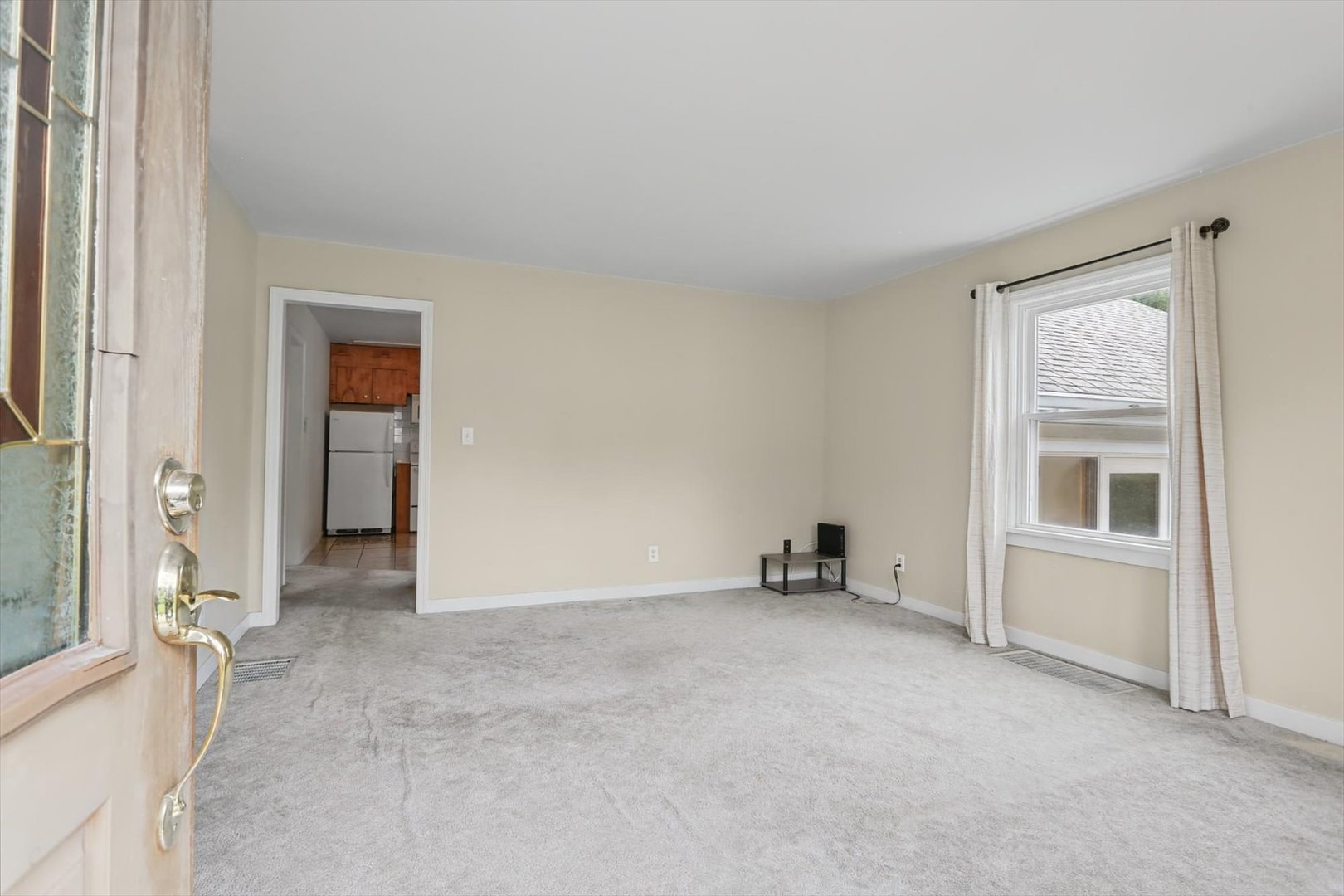 Mostly vacant living room with gray carpet, a curtained window, and a doorway to the kitchen.