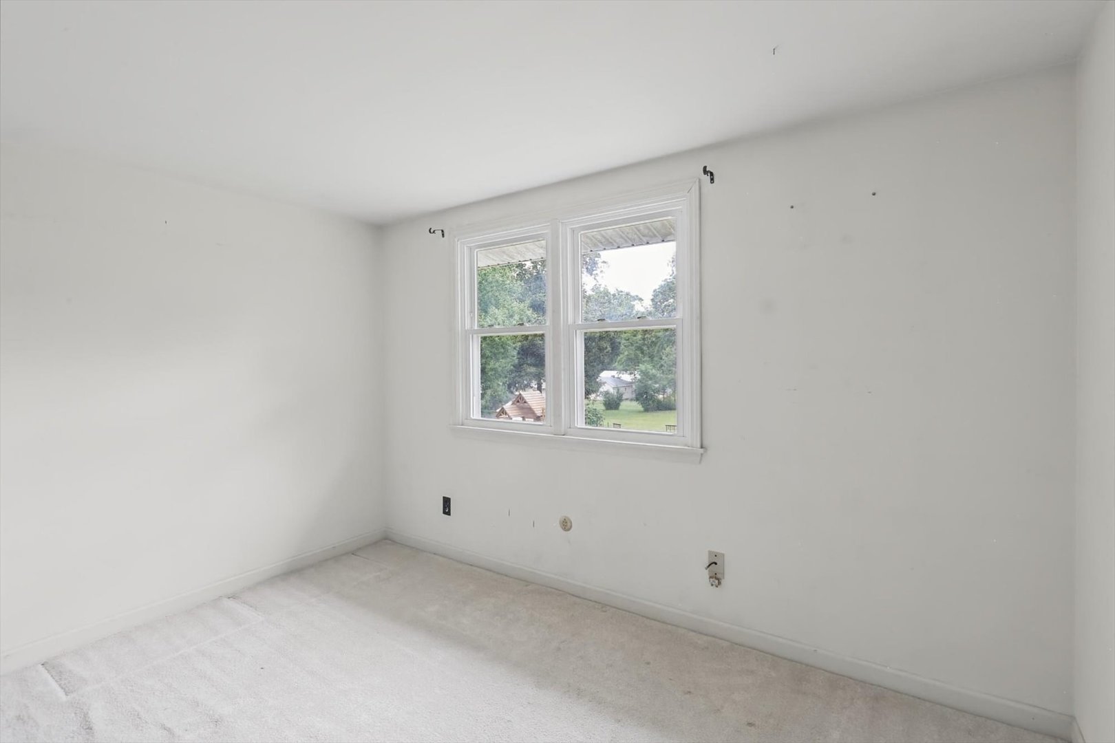 Empty carpeted room with white walls and two windows overlooking a green yard.