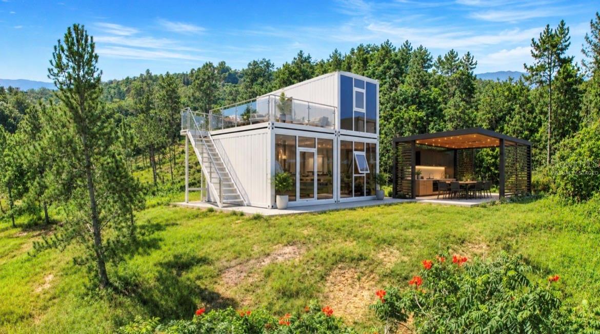 Modern two-story white home with an upper terrace and a lit pavilion in a pine-wooded clearing.