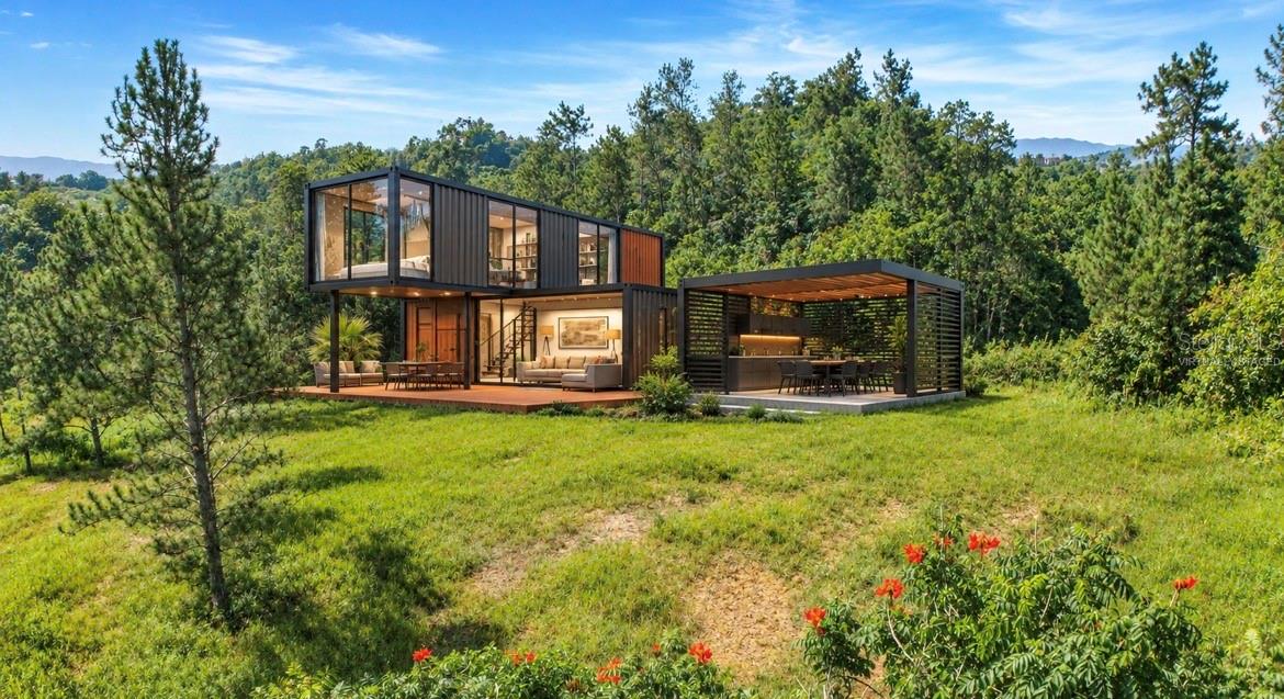 Modern two-story home with large windows, a furnished patio, and a covered dining area in a wooded clearing.