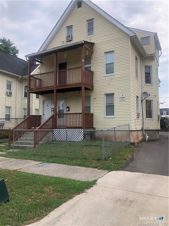 Pale yellow two-story house with stacked brown wooden porches and a fenced front yard.
