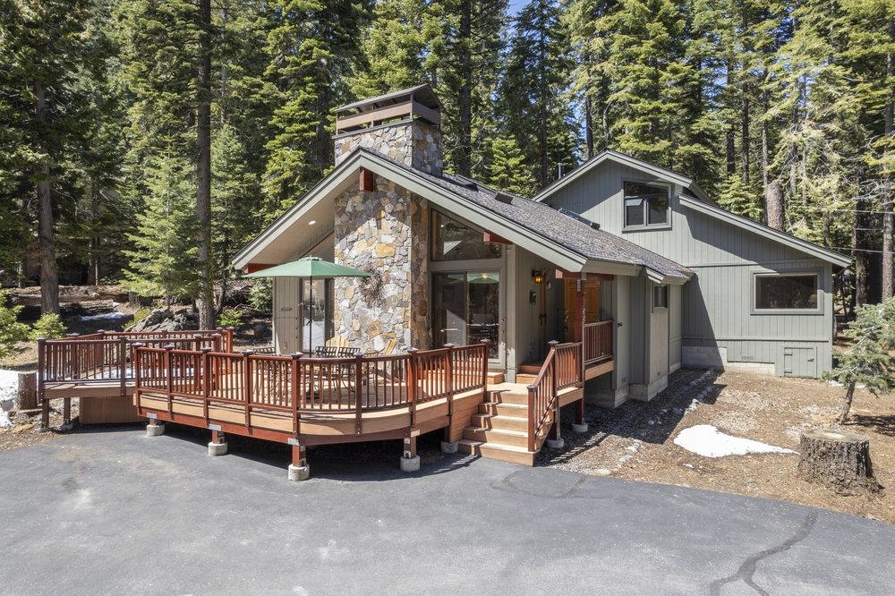 Gray mountain home with a raised redwood deck and stone chimney among pine trees.