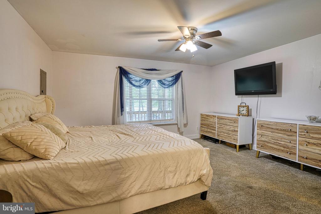 Furnished bedroom with a large beige bed, blue-and-white window curtains, ceiling fan, and wall-mounted television.
