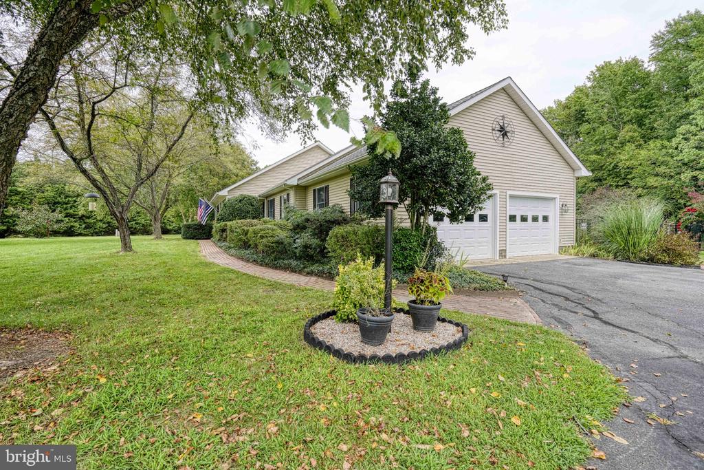 Beige single-story home with double garage doors, mature trees, and a landscaped front lawn.