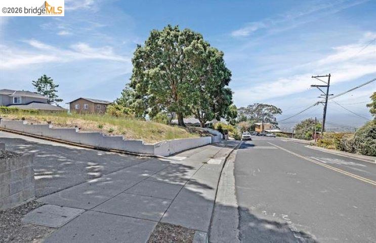 Street and sidewalk beside a sloped landscaped lot, with a large tree and homes in the distance.