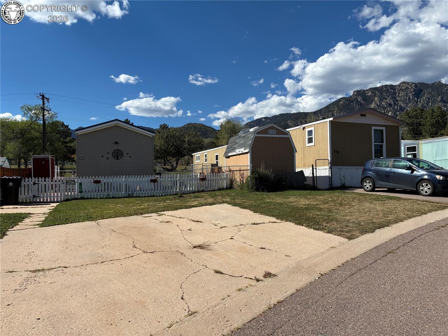 Tan manufactured home marked 119 with a white picket fence and mountains behind it.