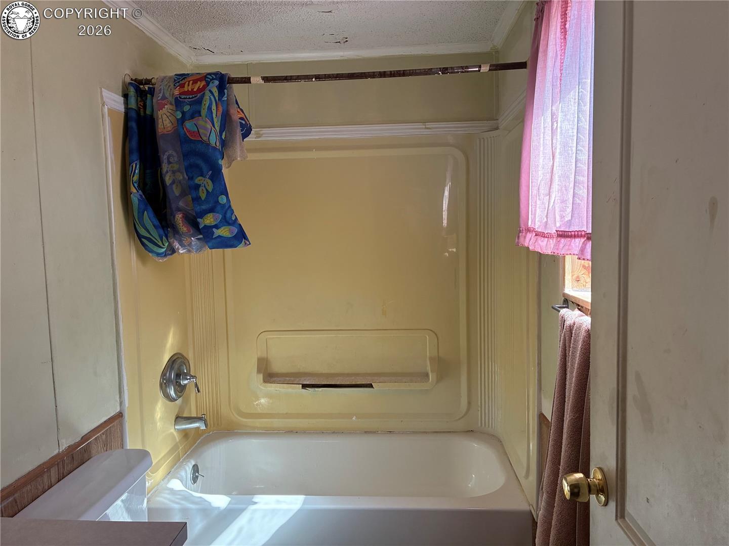 Bathroom bathtub with a yellow surround, built-in shelf, colorful towels, and pink window curtain.