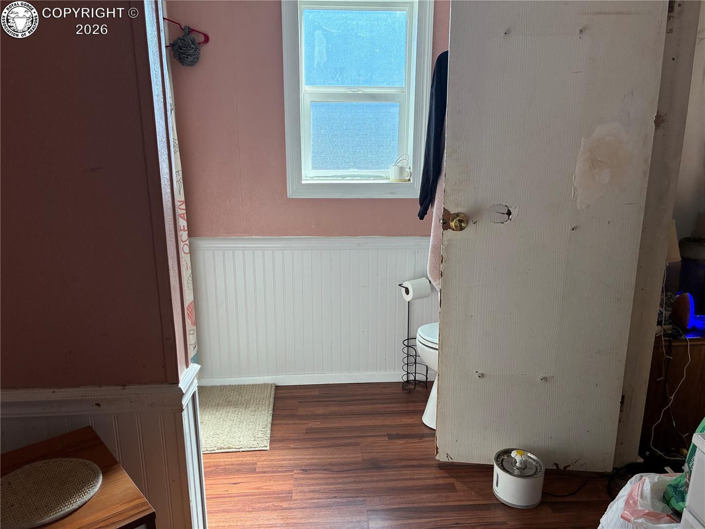 Bathroom with pink walls, white beadboard, a small window, and a damaged door.