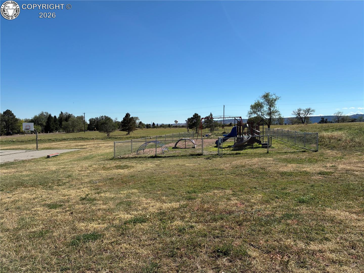Grassy recreation area with a fenced playground, basketball hoop, and distant mountains.