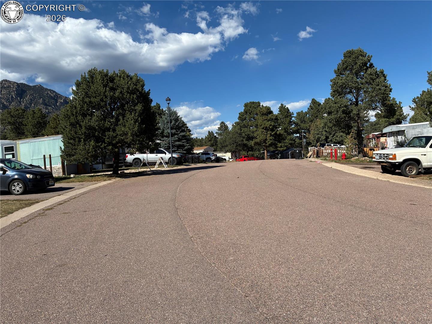 Paved road between manufactured homes, with pine trees and a rocky mountain in the distance.