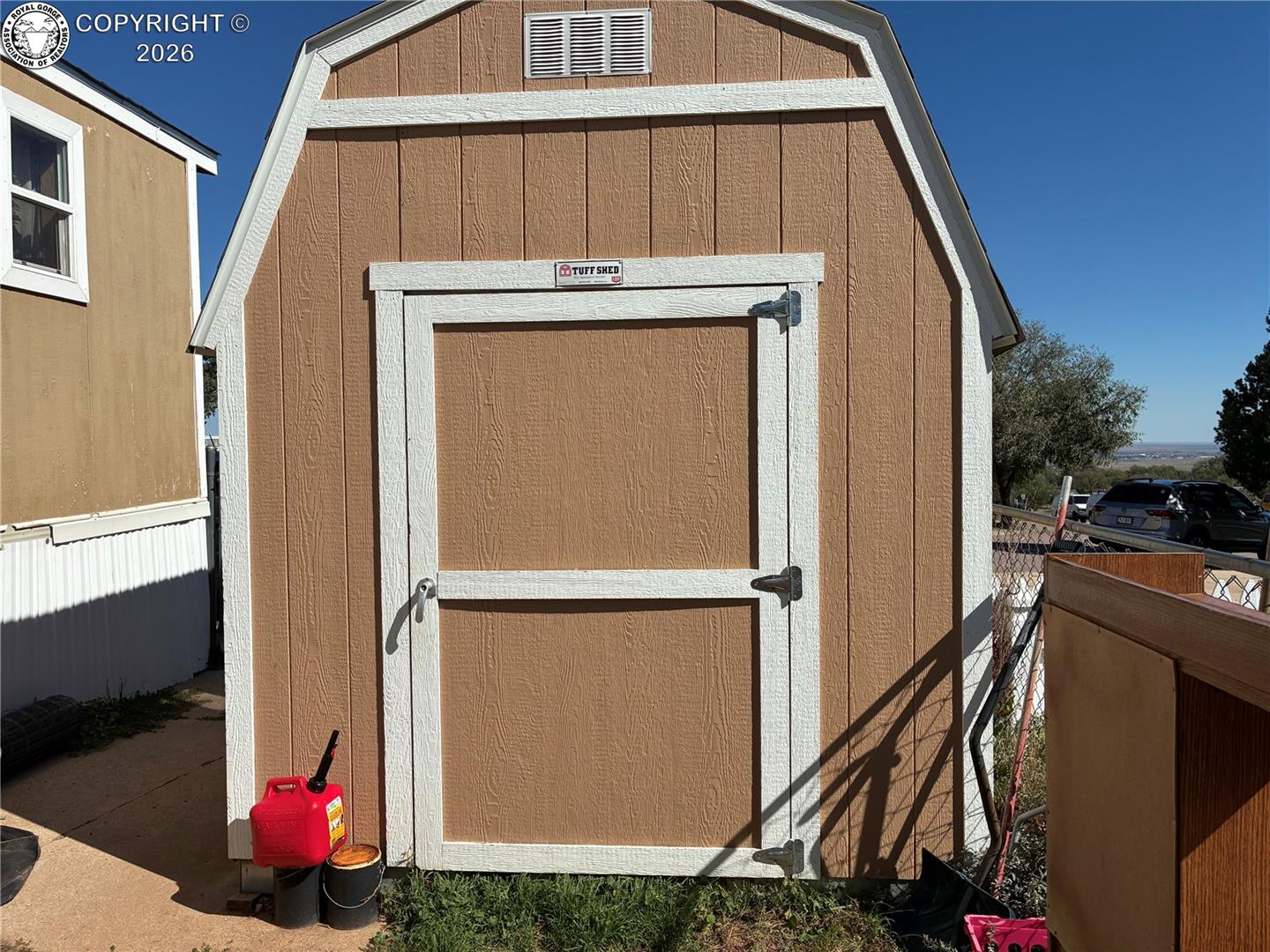 Brown storage shed with white trim and a gambrel roof beside a manufactured home.