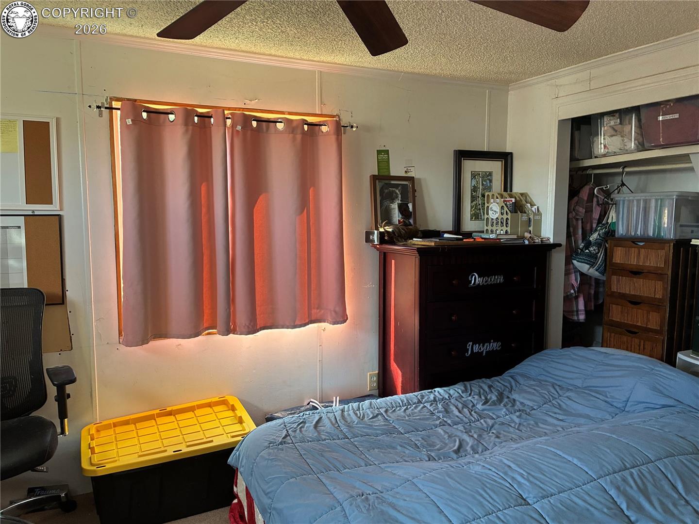 Bedroom with a blue quilted bed, pink curtains, and a dark dresser beside an open storage closet.