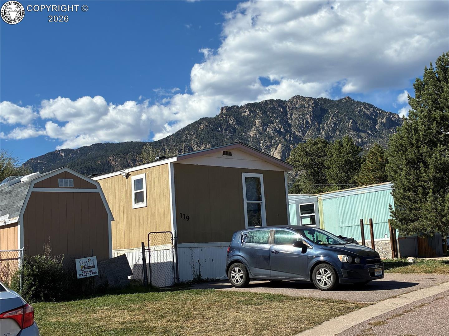 Tan manufactured home marked 119 with a parked car and forested mountain backdrop.