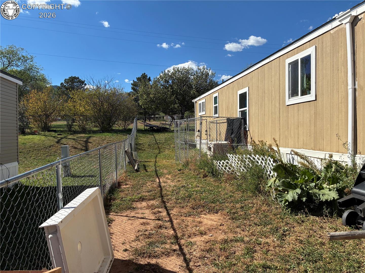 Tan-sided home beside a chain-link fenced yard with patchy grass and shrubs.