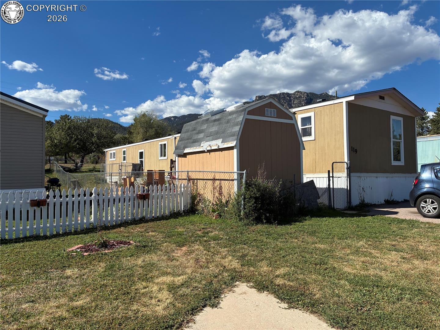 Tan manufactured home marked 119, with a fenced yard, barn-style shed, and mountains behind it.
