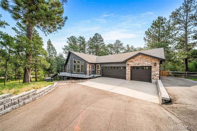 Modern home with stone accents, two garage doors, and a broad driveway among pine trees.