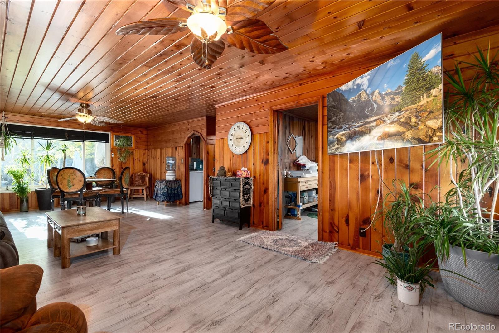 Wood-paneled living room with two ceiling fans, a dining area by the windows, and potted plants.