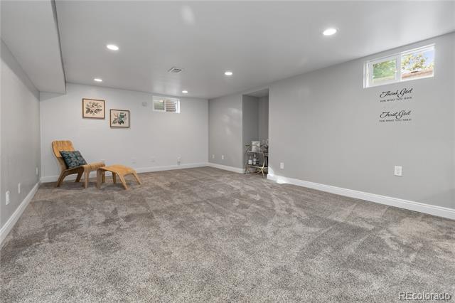 Finished basement room with plush gray carpet, high windows, and a wooden chair with footstool.