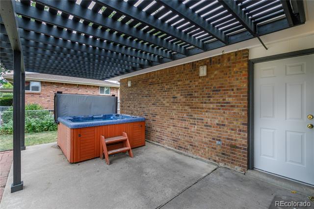 Covered patio with a hot tub beneath a dark slatted pergola beside a brick wall.