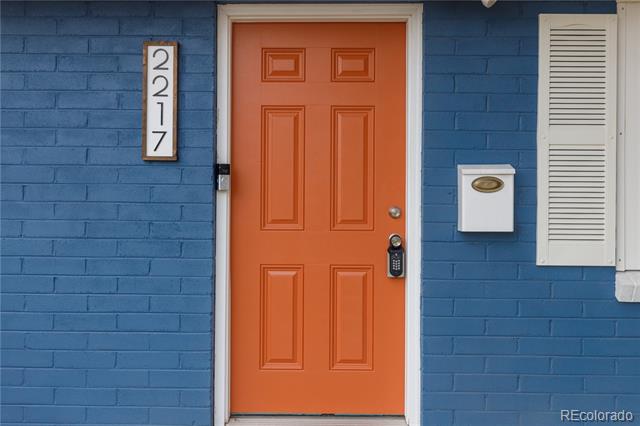 Orange front door set in a blue brick facade, with an address plaque and white shutter.