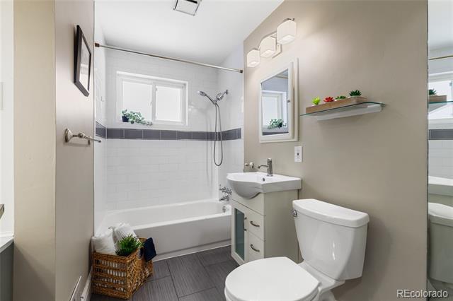 Bathroom with a tub and shower beneath a window, white vanity, toilet, and gray tile floor.