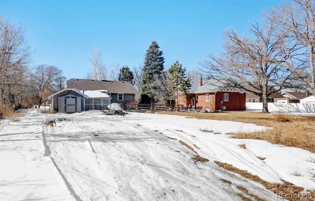 Gray house beyond a snow-covered driveway, with leafless trees and a red building nearby.