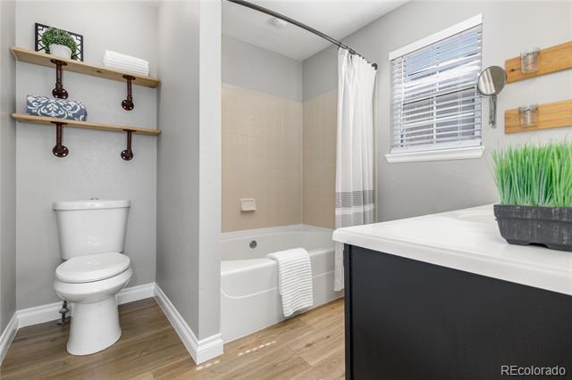 Bathroom with a toilet beneath wooden shelves, a tub and shower curtain, and a dark vanity beside a window.
