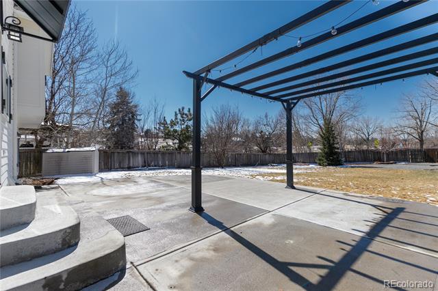 Concrete backyard patio beneath a black pergola, with a fenced yard and leafless trees.