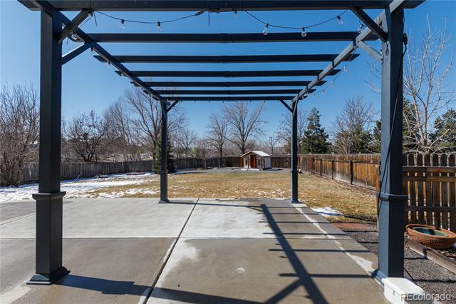 Concrete backyard patio beneath a black pergola, overlooking a fenced lawn with patches of snow.