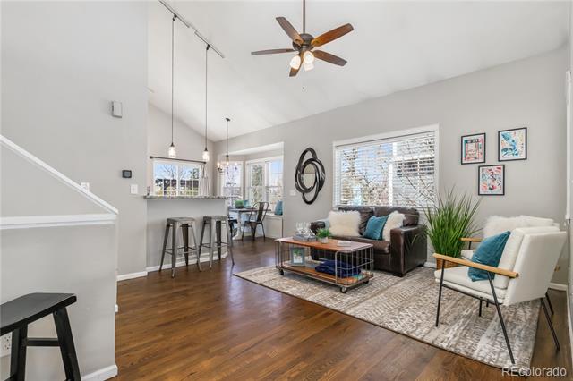 Staged living room with vaulted ceiling, hardwood floors, and seating beside a kitchen bar.