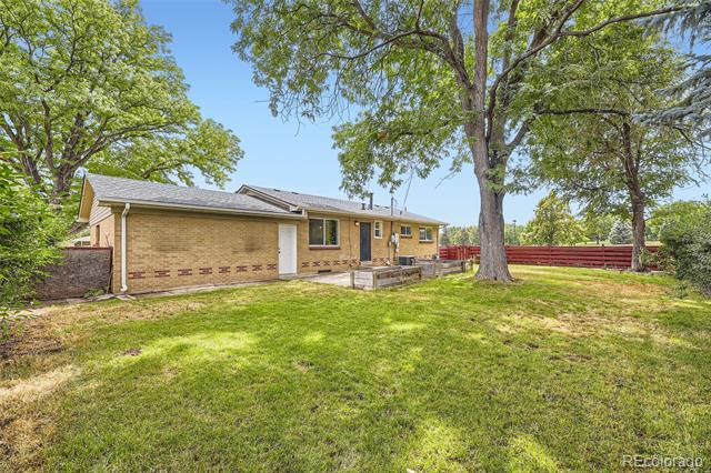 Tan brick ranch house with an attached garage, mature trees, grassy yard, and red fence.