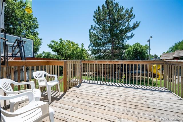 Wooden backyard deck with white chairs, a basketball hoop, and a yellow slide beyond the railing.