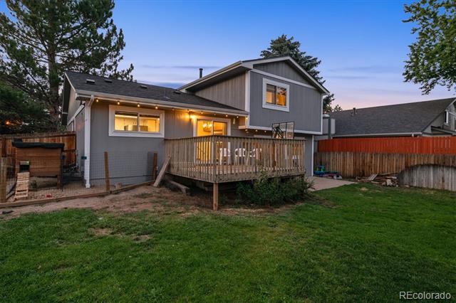 Rear exterior of a gray home with a raised wooden deck and fenced lawn at dusk.