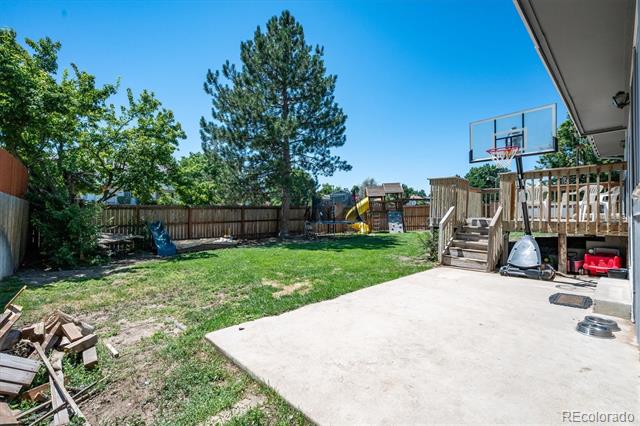 Fenced backyard with a concrete patio, raised deck, basketball hoop, and playset.